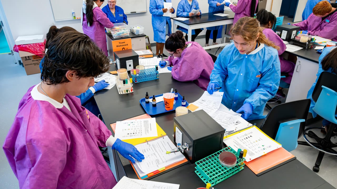 An overhead shot of a lab table with students writing and working, dressed in bright purple and blue scrubs.