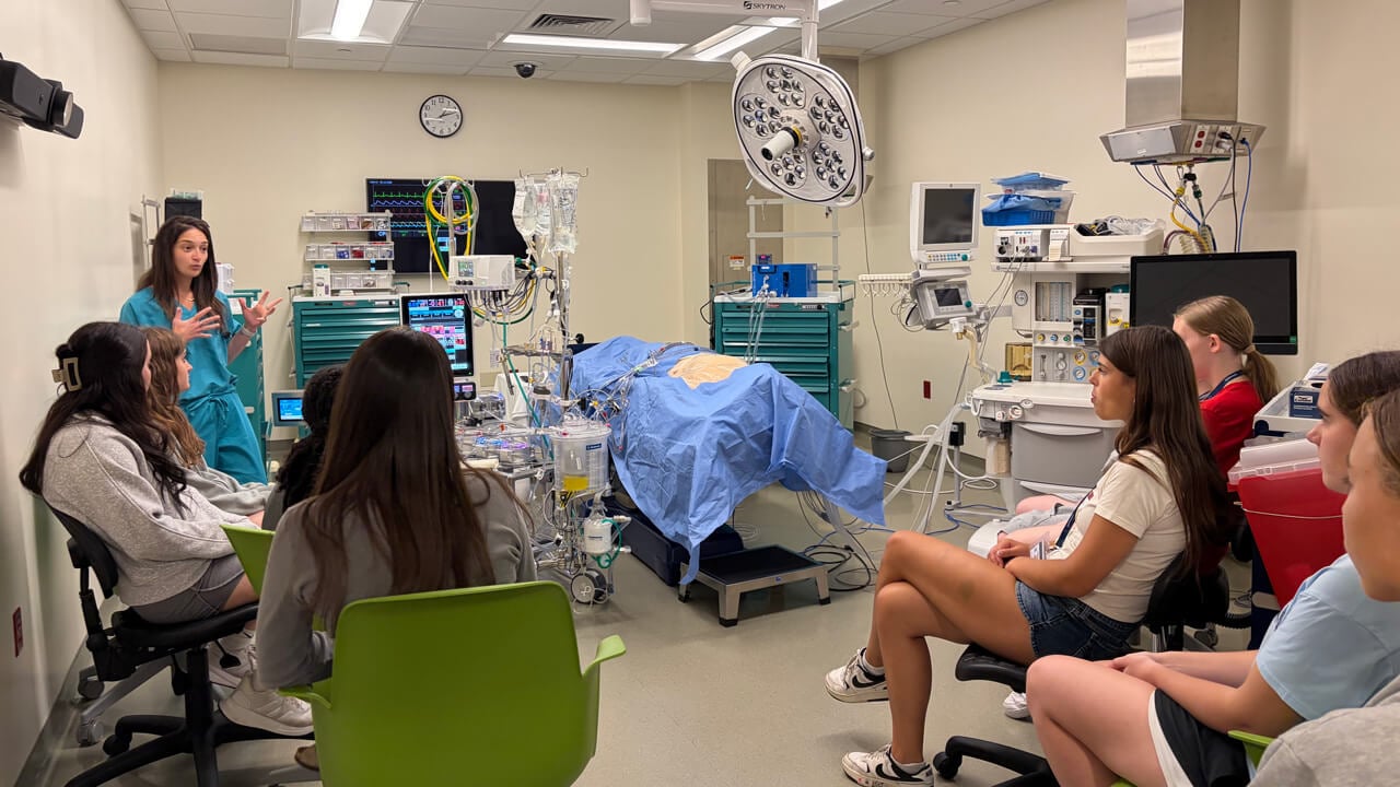 A woman in green scrubs talks to a group of student participants next to a hospital bed simulation.