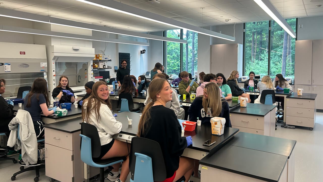 Four lab tables filled with seated students. One of the students closest to the camera is looking at it and smiling.