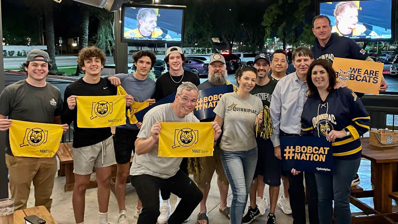 Alumni, students and families stand in their Quinnipiac merchandise at a restaurant watching the national hockey championship game.