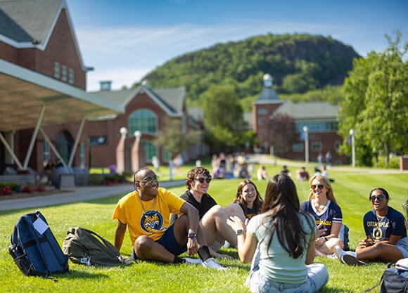 A group of students sits together on a grassy campus lawn, with brick academic buildings and a tree-covered hill in the background.