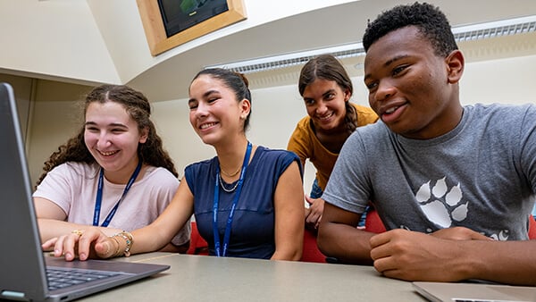 Four students sit and stand together around a laptop at a classroom table, looking at the screen.