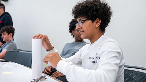 A student works with a white rectangular model at a classroom table while other students sit nearby.