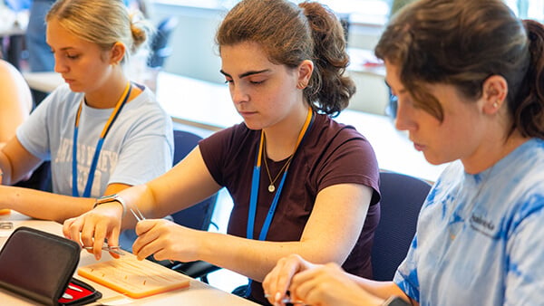 Three students seated at a classroom table use small tools to work with materials during a hands-on activity.