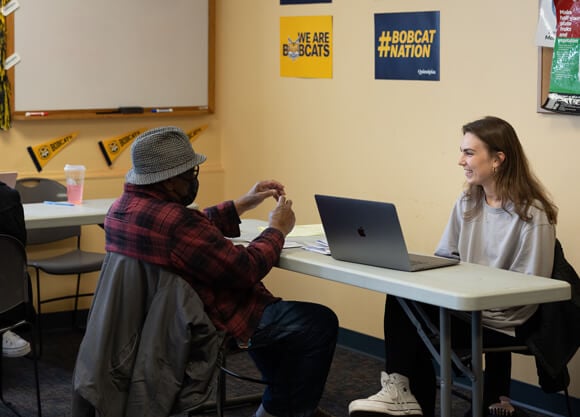 An accounting student sits at a computer and talks with a local resident in the tax clinic
