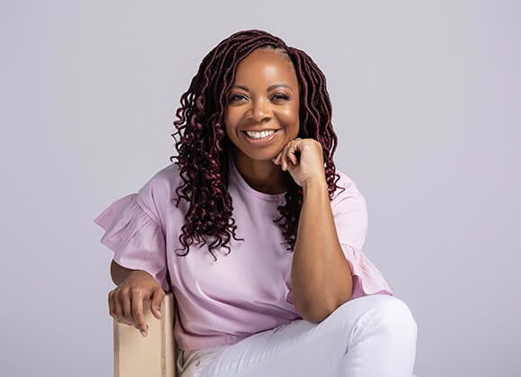 A headshot of Nickia Jackson. She is sitting on a chair with her legs crossed. She is wearing a lavender shirt.