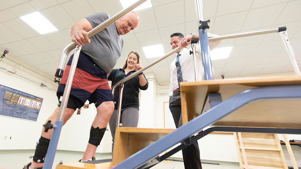 A physical therapy student and professor guide a local elderly patient up a set of stairs in a laboratory setting