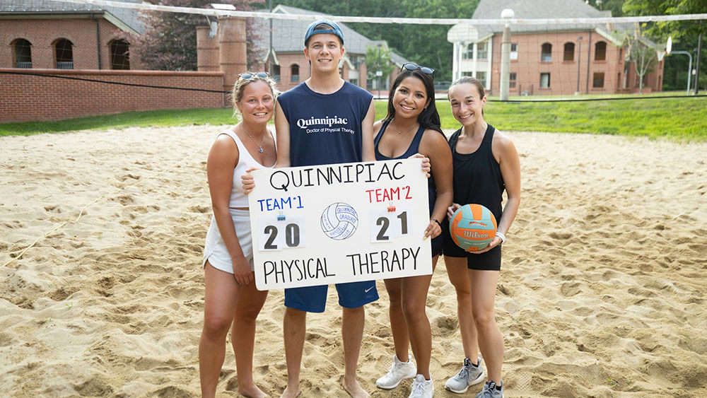 Four physical therapy students pose on a beach volleyball court behind a sign that announces the score of the game