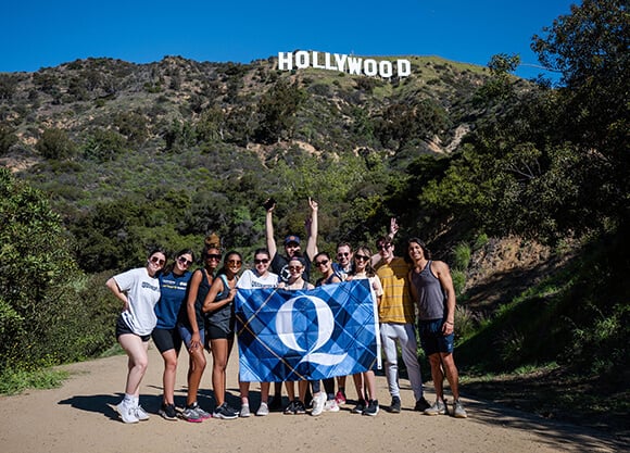 A group of students poses with a Quinnipiac flag on a hiking trail beneath the Hollywood sign in Los Angeles.