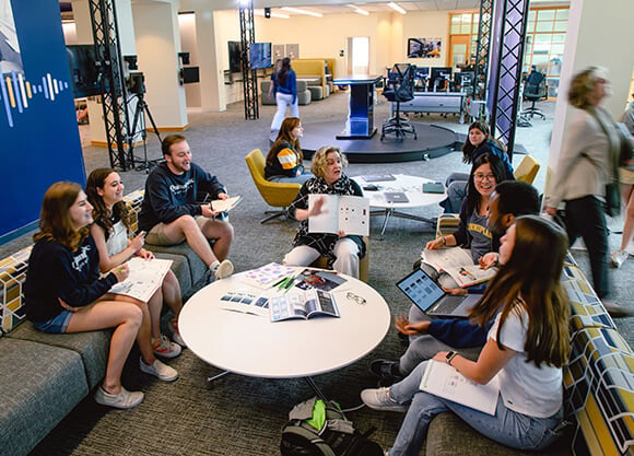 A group of students works with printed materials and tablets around tables in a collaborative learning space with a professor leading a discussion