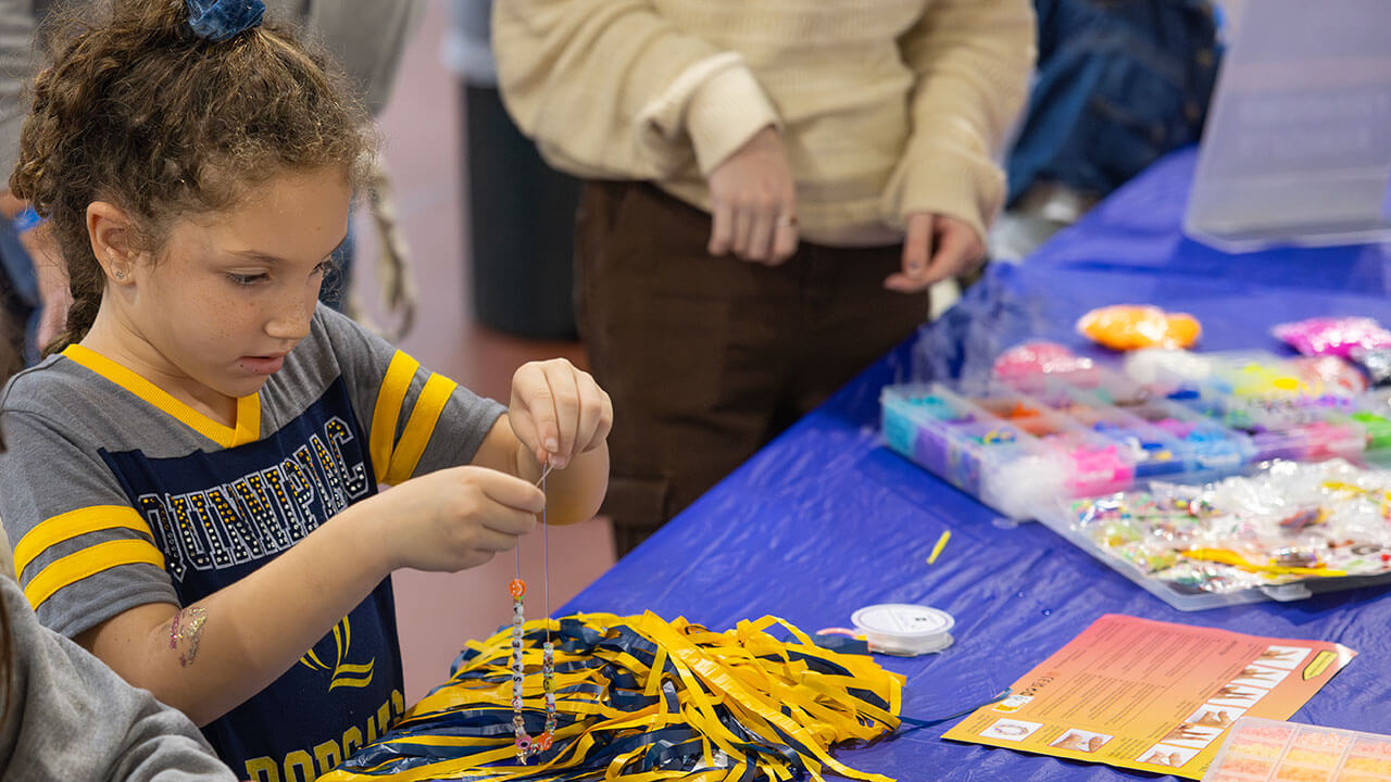 A young girl in Quinnipiac spirit wear focuses in as she beads a bracelet