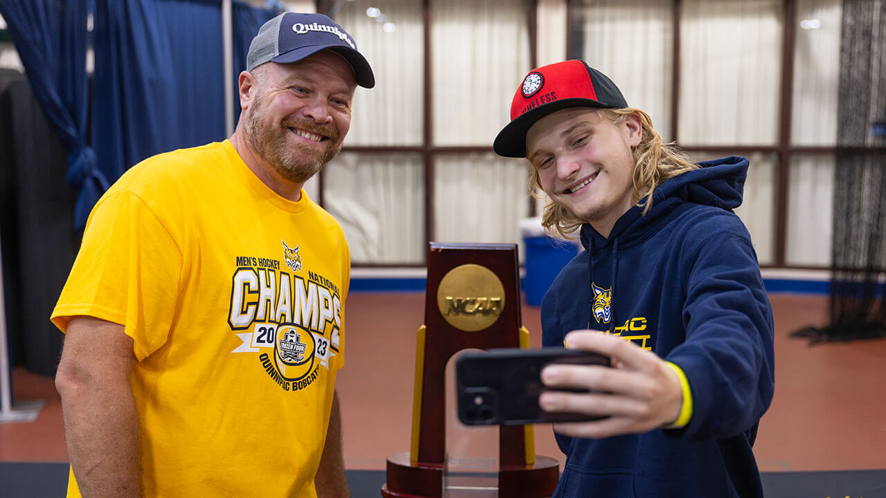 Two guys in Quinnipiac spirit wear take a selfie as they smile in front of the NCAA National Championship trophy