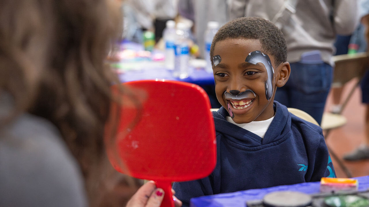 A boy with face paint smiles into a mirror