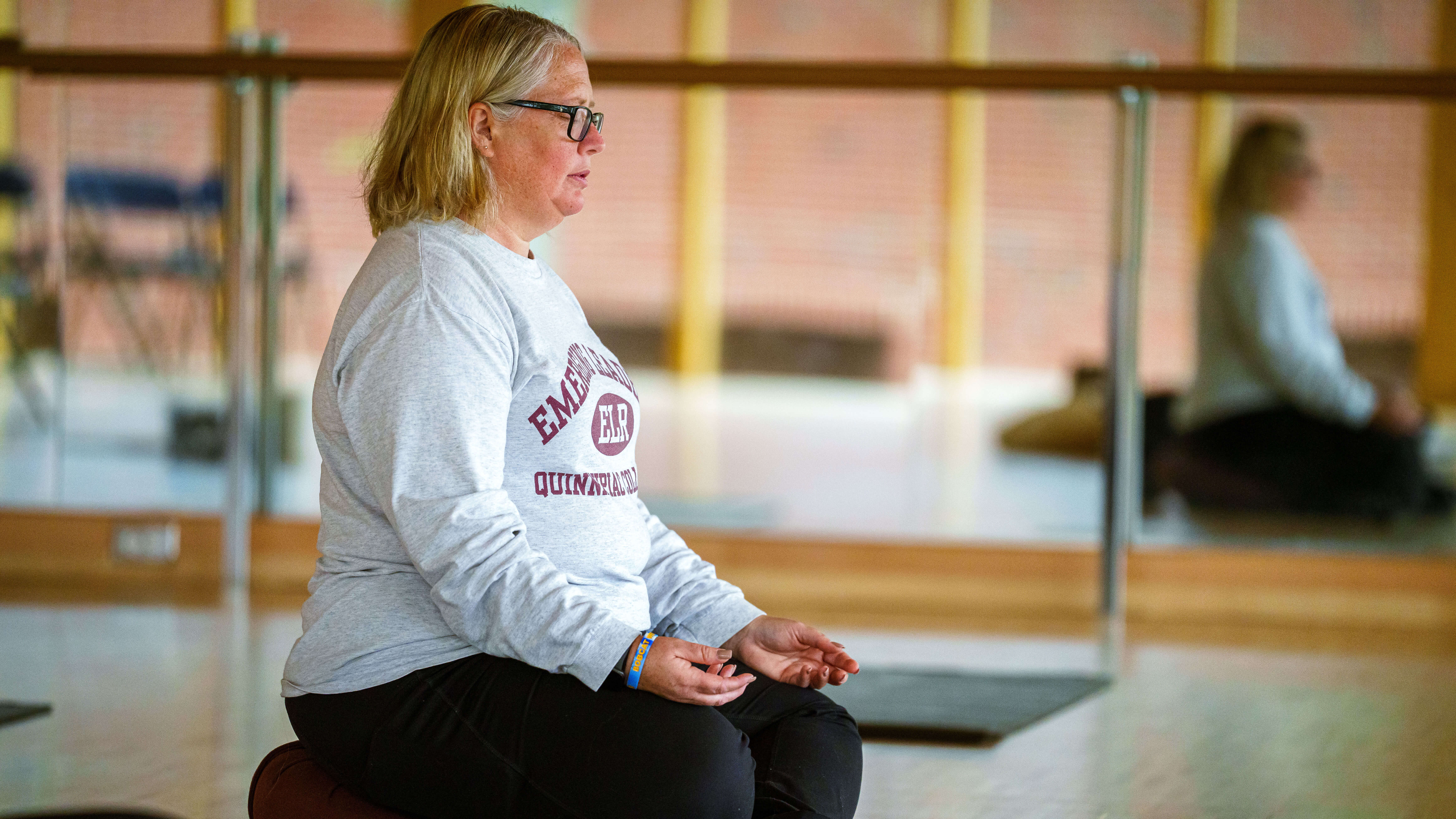 A woman with glasses sits on her knees in a yoga position.