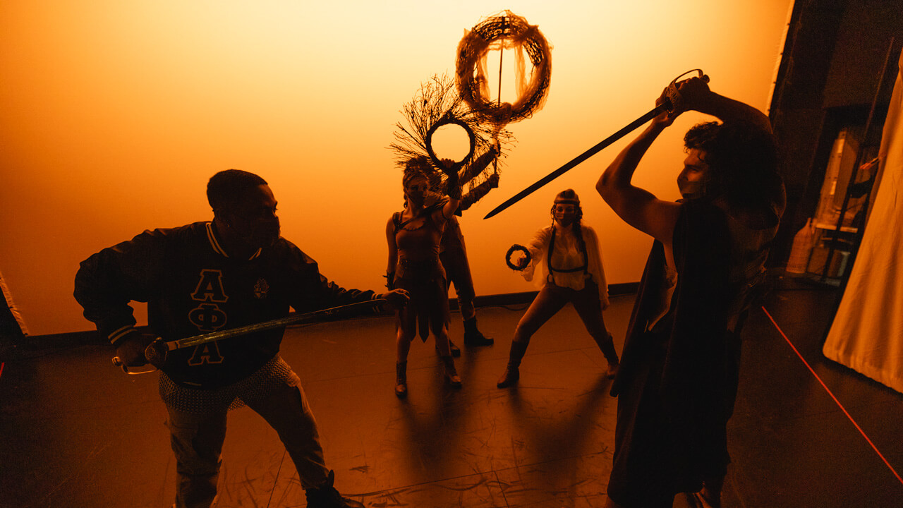 A group of student actors on stage for the production with orange lighting background, An Iliad