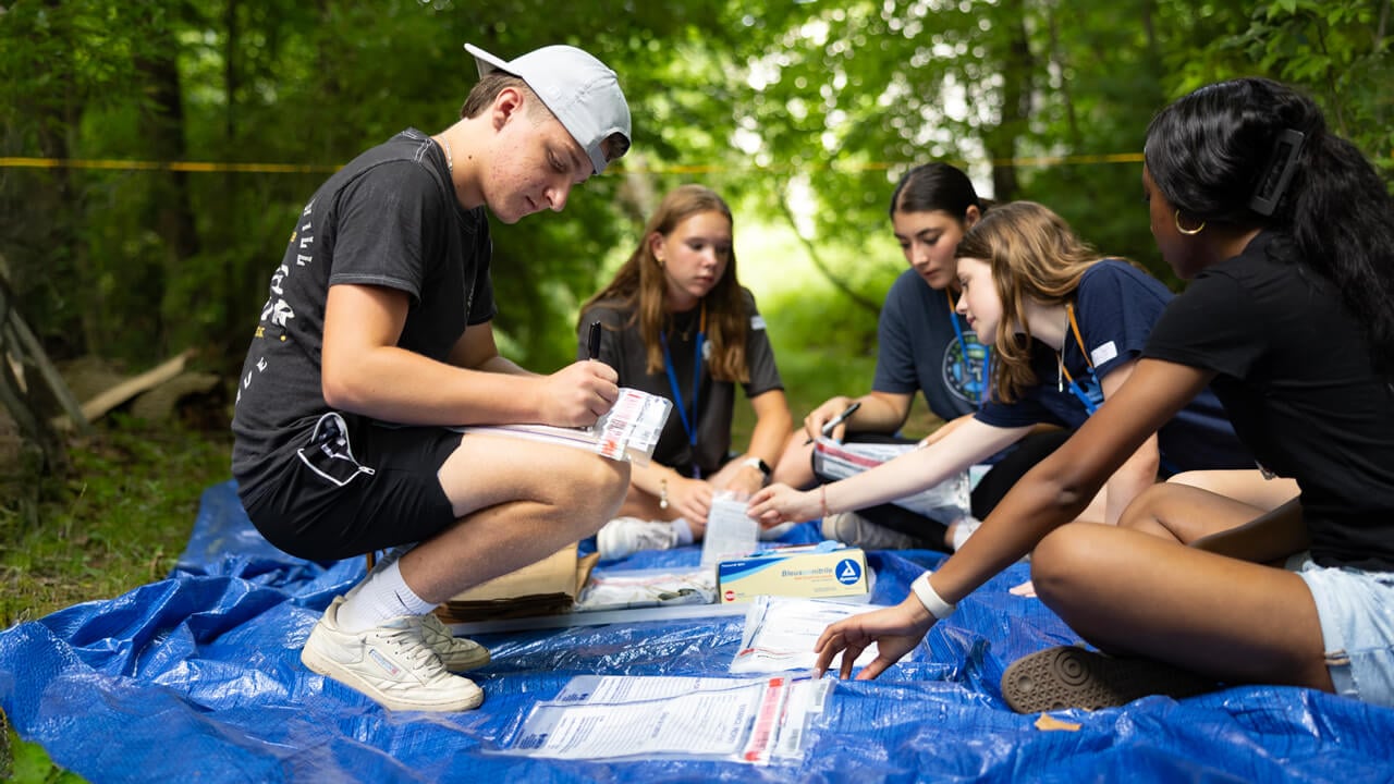A group of high school students sit together on a blue tarp outdoors, labeling and organizing evidence bags as part of a hands-on forensic science workshop at Quinnipiac University.