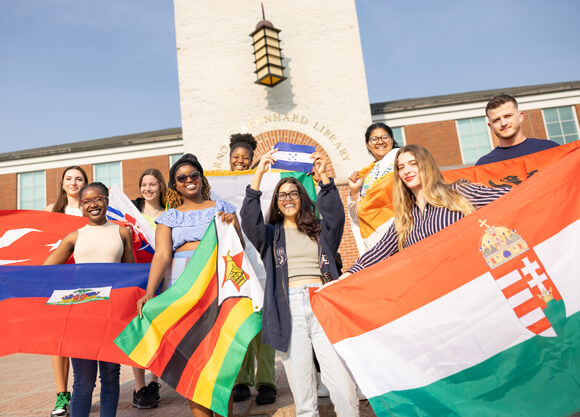 9 international students hold up their country's flag on the steps of the Quinnipiac library
