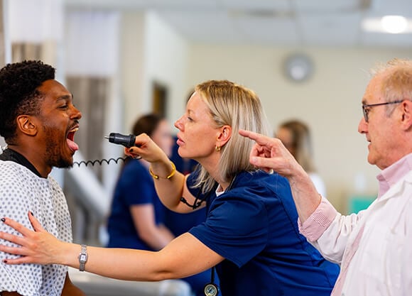 A nursing student performs a throat examination on a standardized patient while an instructor observes and provides guidance during a clinical training exercise.