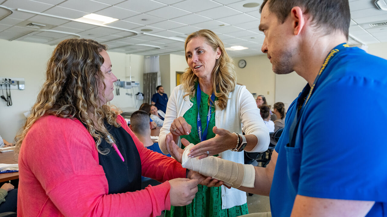 A nursing student practices clinical skills on another student during a healthcare training session as an instructor demonstrates proper arm bandaging techniques in a classroom setting.