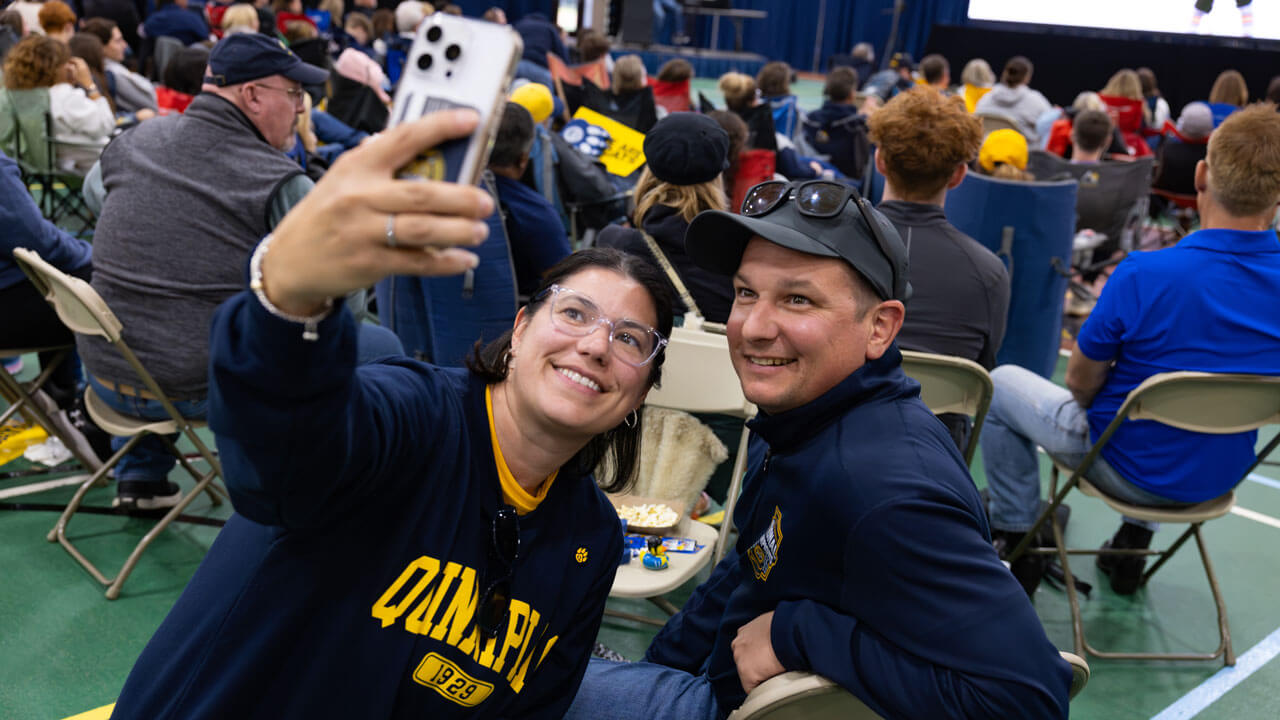 Two individuals pose for a selfie amongst a crowd of people sitting in chairs