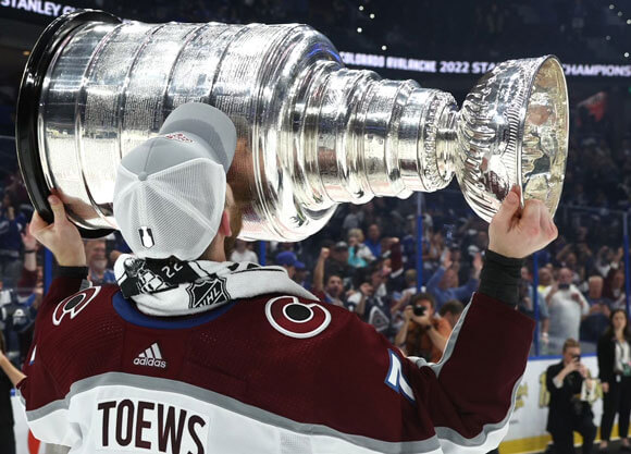 Devon Toews kisses the large Stanley Cup trophy with his back to the camera and his name on his jersey showing.