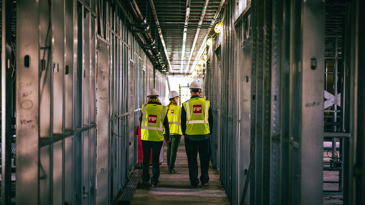 Construction workers walk through an unfinished building wearing bright vests and hats.