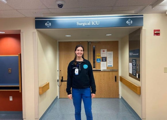 Danielle Petrovich stands in front of a surgical ICU unit in her nurse uniform.