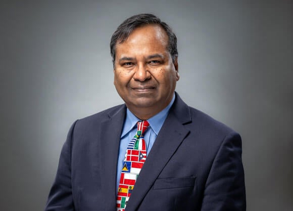 Professor Mohammad Elahee posing for a headshot on a gray background wearing a tie with many different country flags on it.