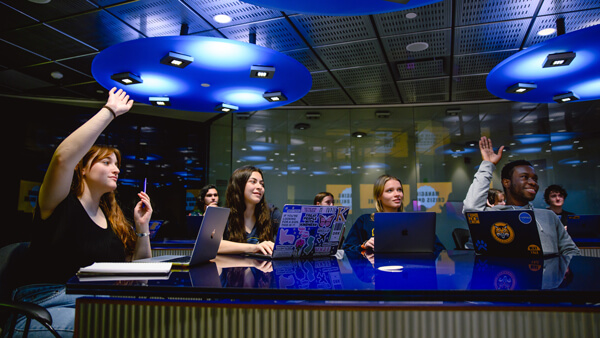 Four international students sit at a table and raise their hands in a communications class