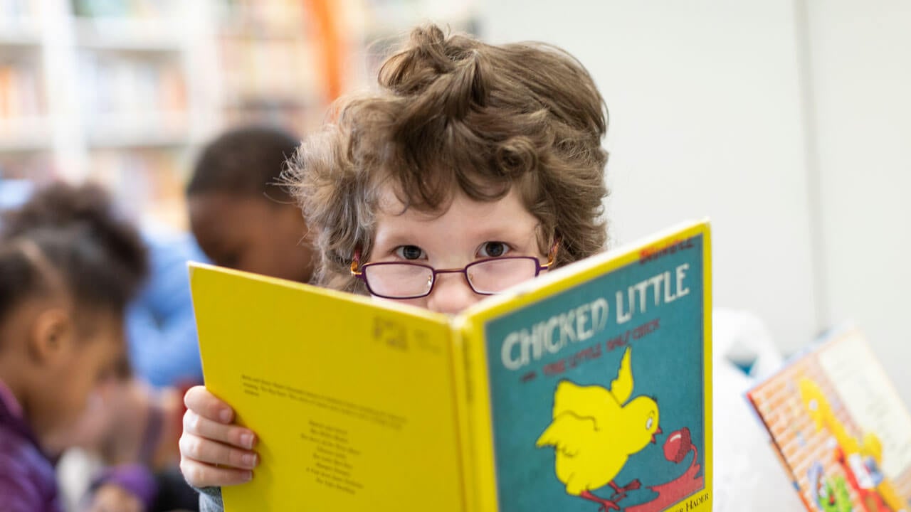 The top of a child's head wearing glasses is visible above an open bright yellow book.