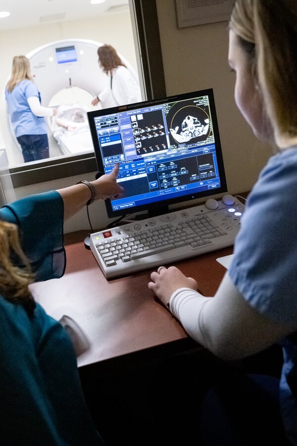 Student viewing imaging results on a computer while a patient goes through on MRI machine