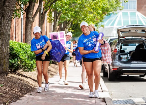Four students in blue shirts carry items to a residence hall during movein.