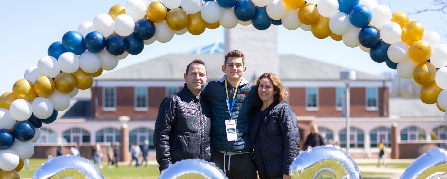 An admitted student and his parents smile under a balloon archway on the quad