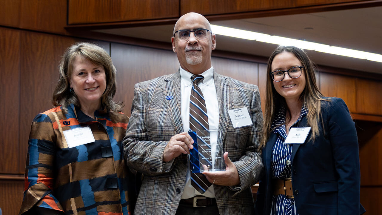 An awardee smiles holding a trophy next to two other professionals at the inaugural alumni association awards.