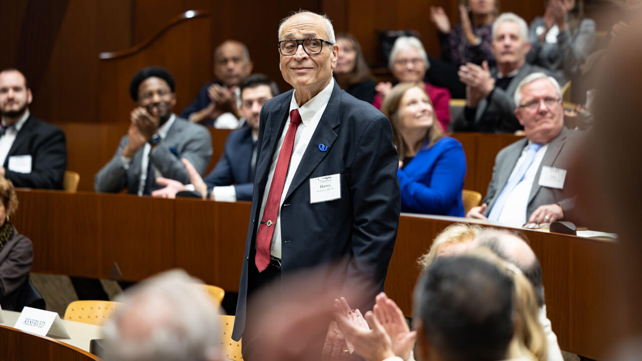 An awardee wearing a suit and red tie stands and smiles as audience members clap.