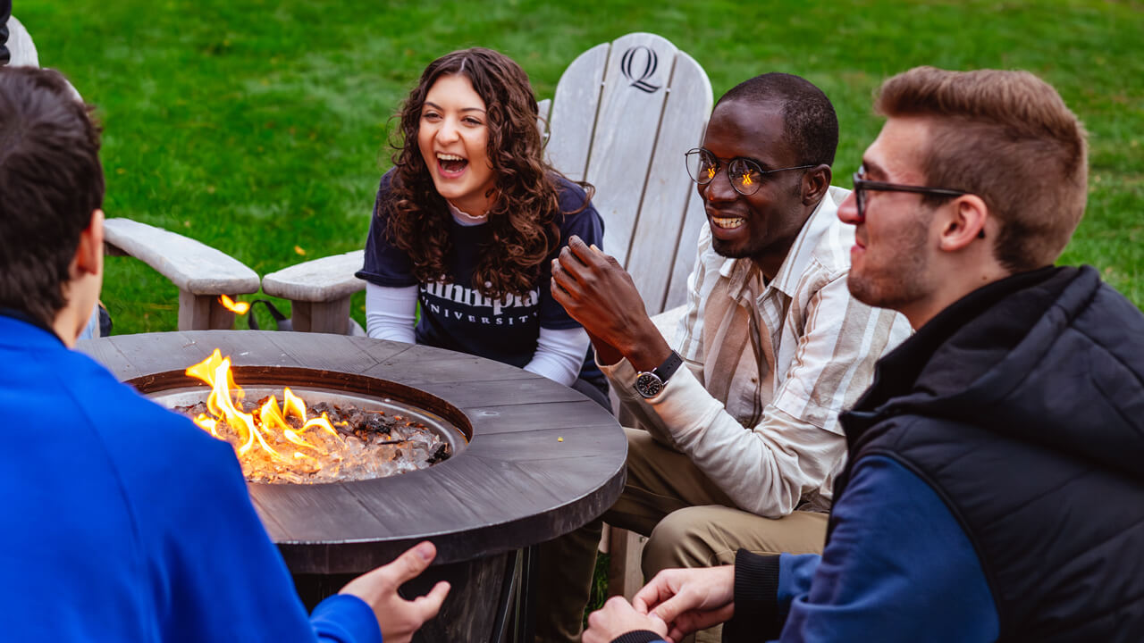 Students sit, talk and laugh on Adirondack chairs around a fire pit on the Quinnipiac quad