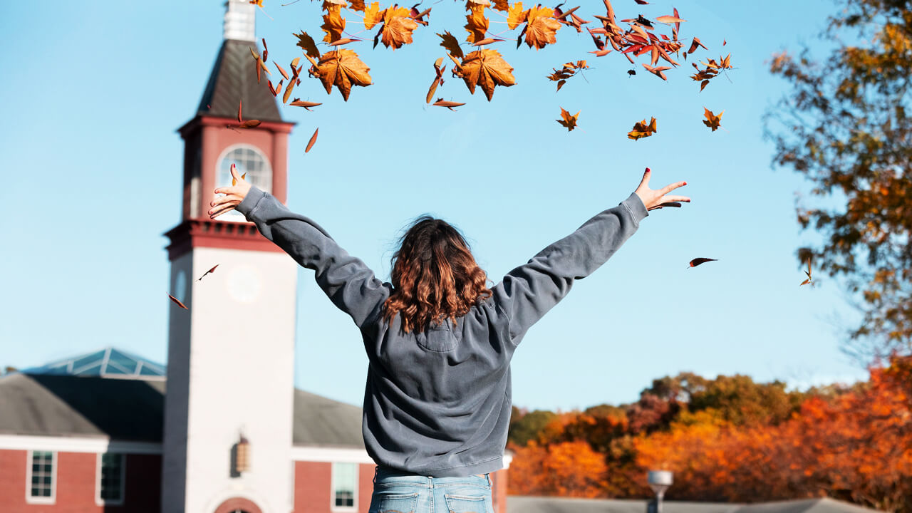 A student throws autumn leaves in the air in front of the Quinnipiac clock tower