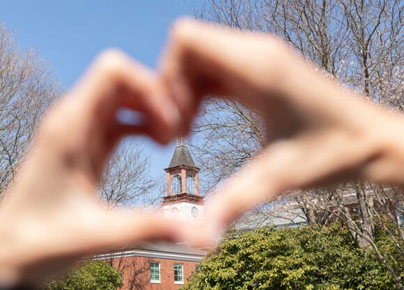 An admitted student forms her hand into a heart with the Quinnipiac clocktower in the background