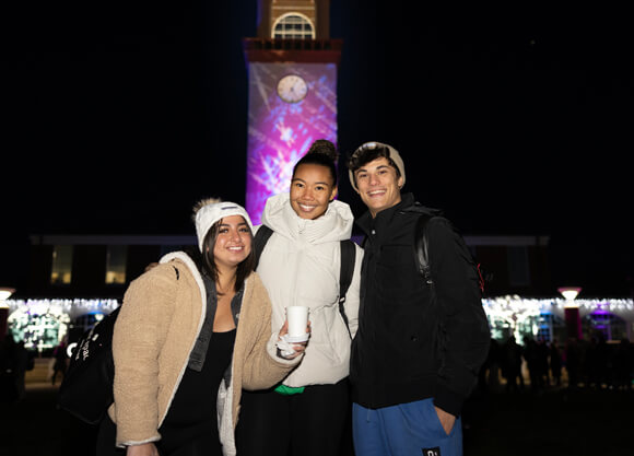 Three students in winter coats pose in front of the library illuminated with holiday lights
