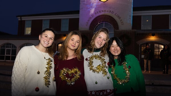 Four students wear festive sweaters that spell I heart Q U in front of the illuminated clocktower