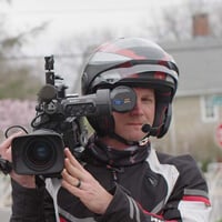 Matt Andrew wears a helmet and racing jacket and looks through the lens of a camera