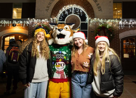 Three students in winter hats pose with Boomer by the lighted clocktower