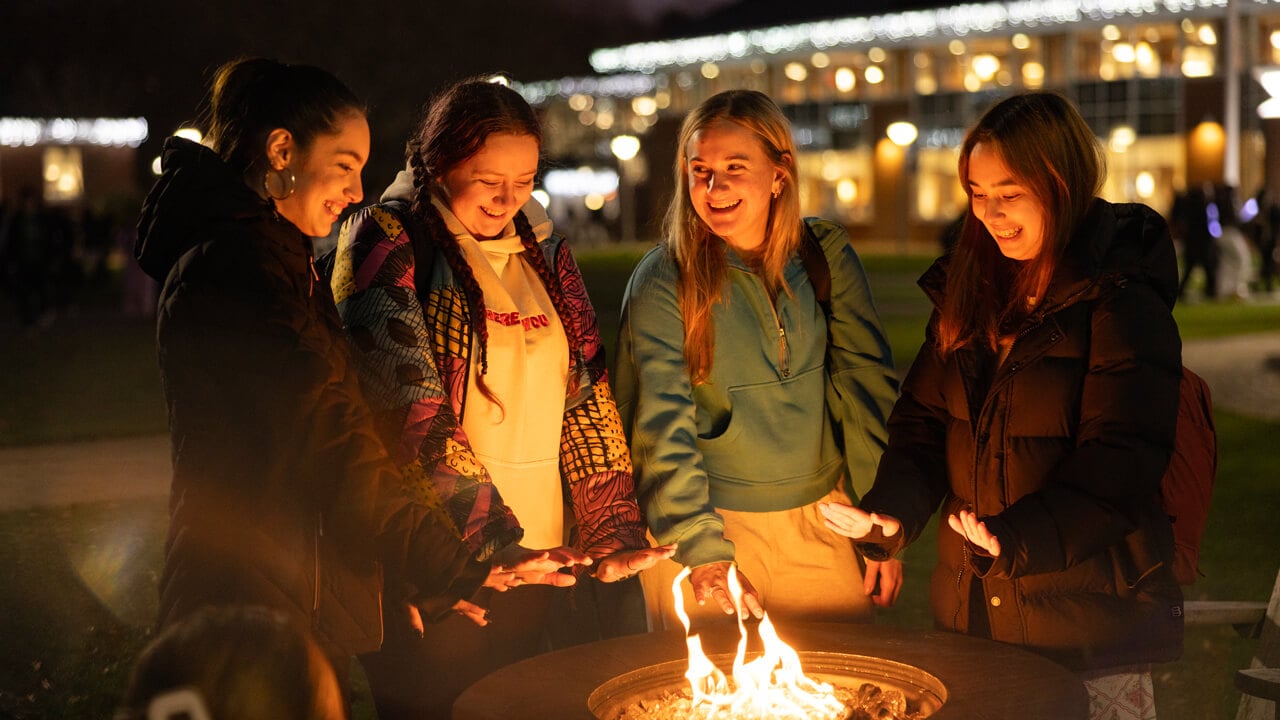 Four students talk and warm their hands at an outdoor fire on the Quad with holiday lights
