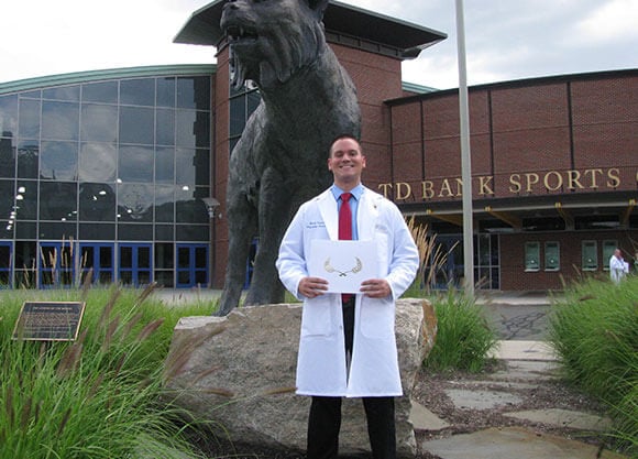 Mark Turczak wears his lab coat and holds his diploma next to the bobcat statue