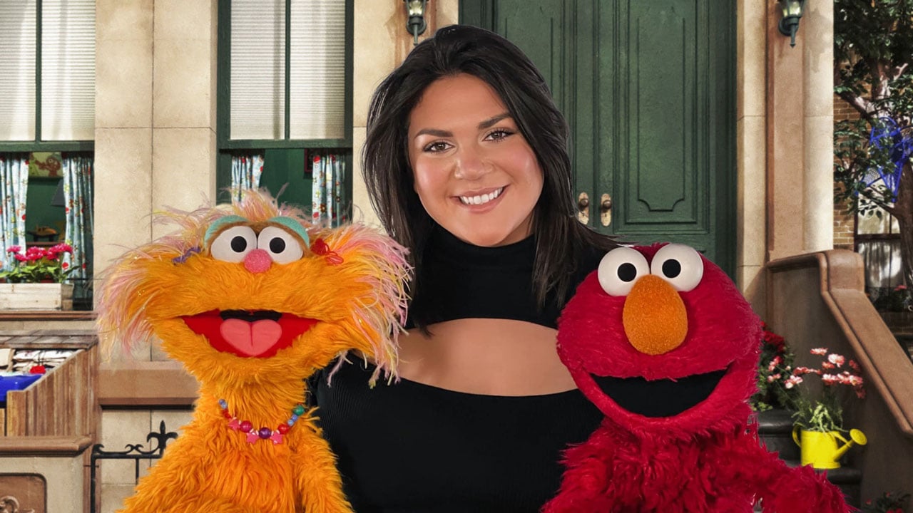 Christina Vittas smiles on Sesame Street between Elmo and an orange muppet character.