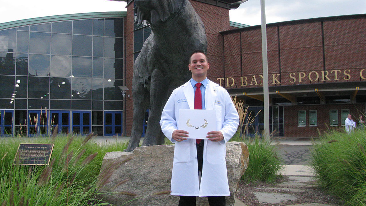 Mark Turczak wears his lab coat and holds his diploma next to the bobcat statue