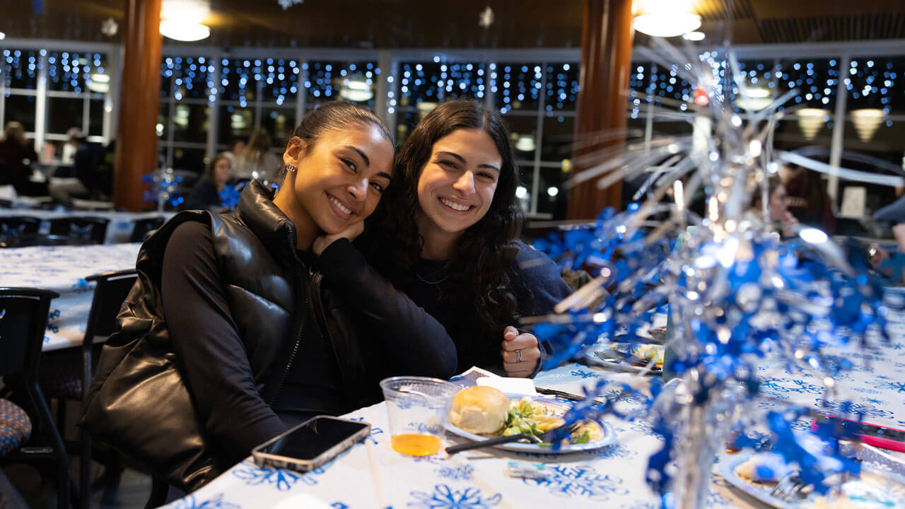 Two girls sit together and smile at undergraduate holiday dinner