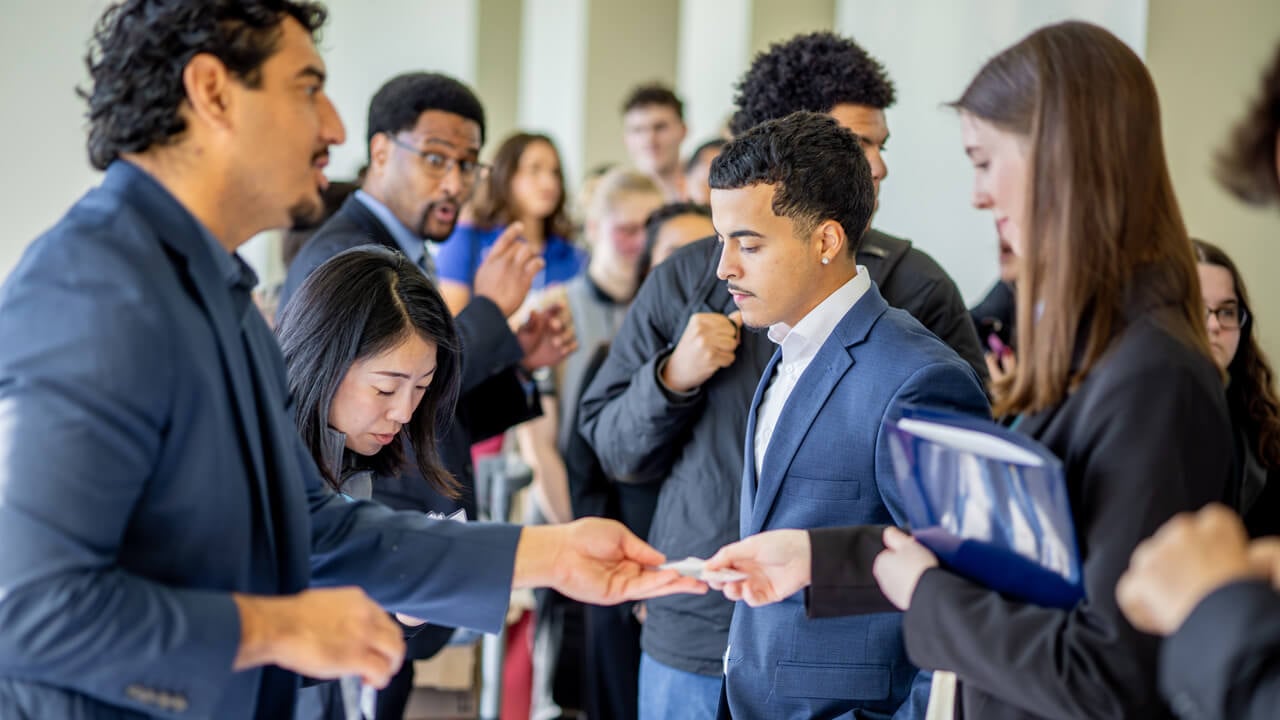 An exhibitor hands a business card to a student in a busy event space.