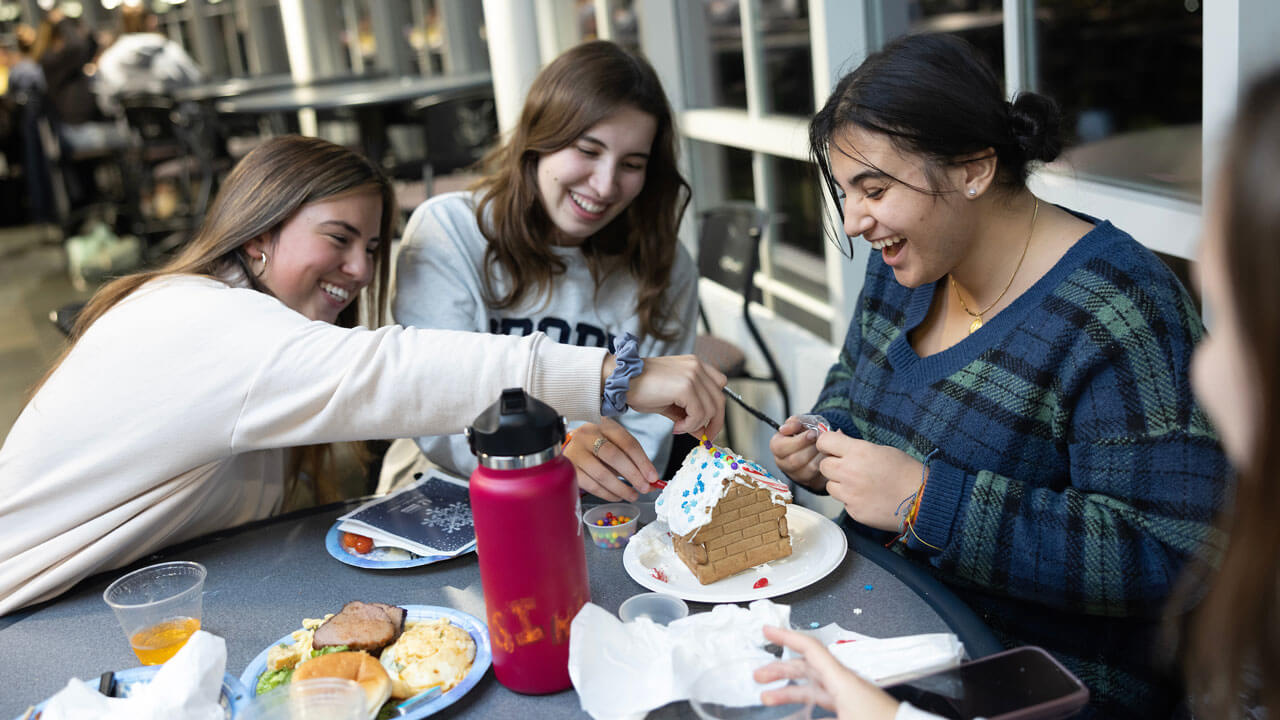 Three students smile and laugh as they deocrate a ginger bread house together at undergraduate holiday dinner.
