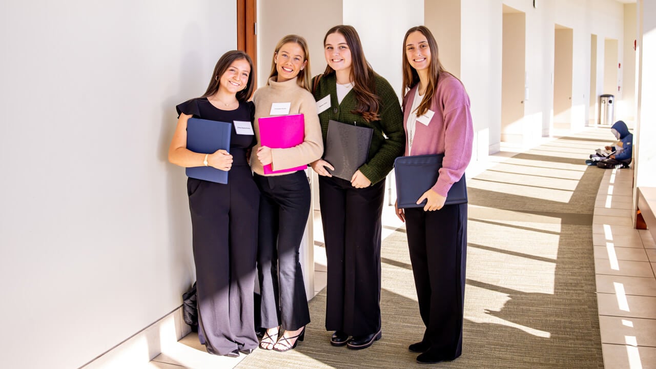 Four female students smiling in a hallway holding binders.